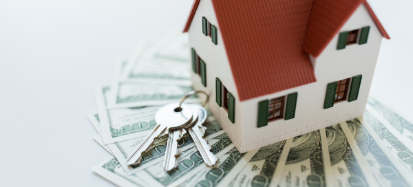 A model house sitting on fanned-out hundred-dollar bills with a set of house keys, representing modular home prices and financing in Maryland.